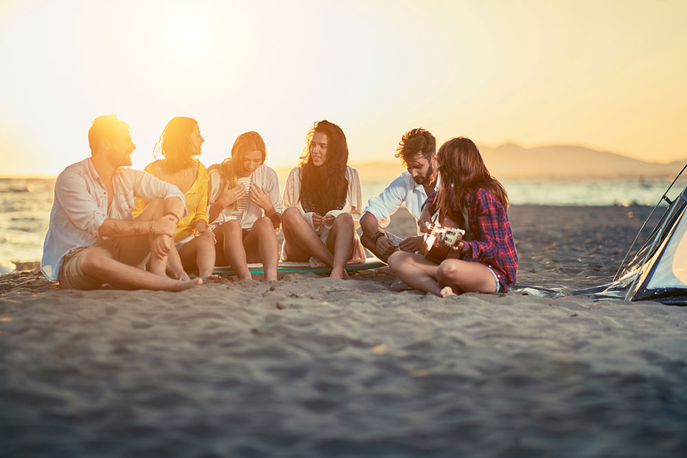 Group of young friends with guitar at beach. friends relaxing on sand at beach with guitar and singing on summer sunset