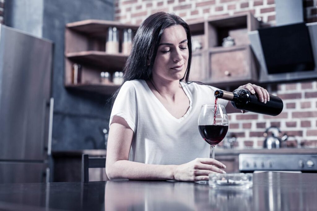 Sad cheerless brunette woman sitting at the table and holding a bottle while pouring wine
